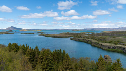 Tranquil summer view of Lake Myvatn in northern Iceland, with calm waters, volcanic terrain, and lush greenery under a bright blue sky.
