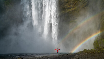 Unidentified woman stands beneath the powerful Skogafoss waterfall in Iceland, framed by mist and a vivid rainbow