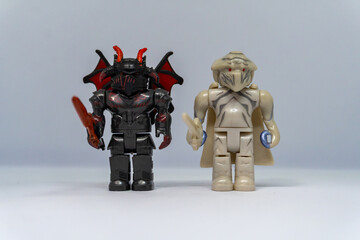 Two block figures, one dark with red horns and wings holding a sword, the other light-colored with...