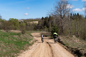 Two cyclists and a dog enjoy a sunny ride along a dirt road, surrounded by lush greenery and a...