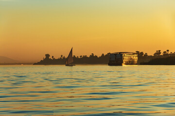 Traditional sailboat and cruise ship on the Nile River at sunset in Luxor, Egypt with golden sky...