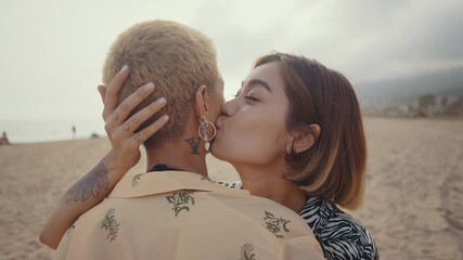 Passionate lesbian couple sharing tender moment while embracing and kissing on sandy beach during romantic sunset, expressing deep love and connection