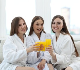 Happy women in bathrobes with glasses of orange juice on sofa in spa salon