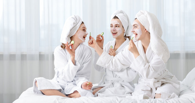 Happy women with facial masks and cucumber slices in bathrobes after spa procedures on bed indoors
