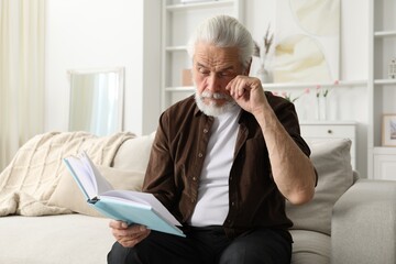 Senior man with vision problem reading book on sofa at home