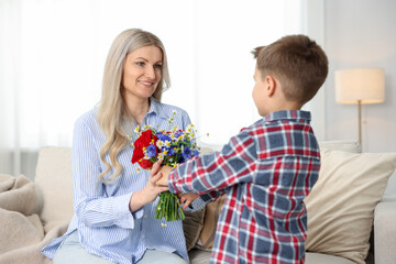 Little boy presenting bouquet of beautiful flowers to his smiling mother on sofa at home