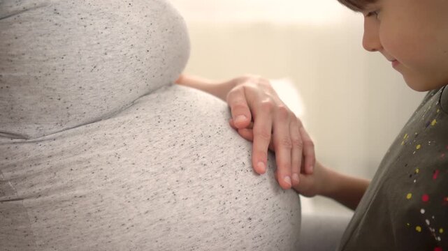 Little brother strokes his mother's pregnant belly. The boy smiles, feeling movements of baby. Happy moments during pregnancy. Motherhood and parenting concept. Happy family expecting for baby.