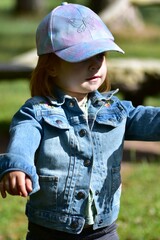 Toddler girl in pastel butterfly cap and denim jacket standing outdoors with arms open. Scene symbolizes openness, joy, and freedom, capturing essence of playful childhood in nature on a sunny day.