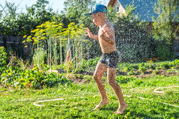 Joyful boy in a cap enjoys splashing water from a hose, playfully cleaning himself in a vibrant garden under bright sunshine