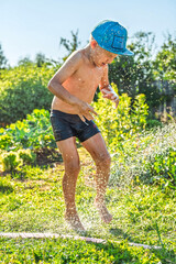 Joyful boy in a cap enjoys splashing water from a hose, playfully cleaning himself in a vibrant garden under bright sunshine