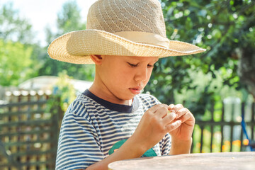 Small boy in a straw hat focuses on shelling beans while sitting at a table outside, surrounded by greenery