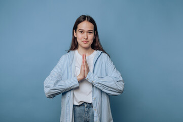 Young woman with long hair expresses gratitude with hands in prayer pose against a blue background...