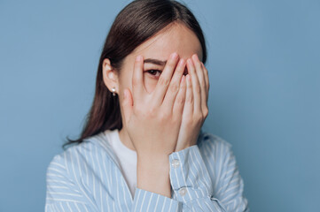 Young woman covers her face with hands against a blue background, expressing shyness and vulnerability