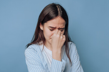 Woman expresses distress in front of a solid blue background while holding her nose with her eyes...