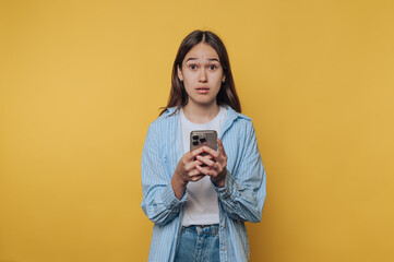 Young woman holding smartphone with surprised expression in front of yellow background