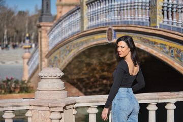 Young, beautiful, dark-haired woman in a black long-sleeved T-shirt and jeans, with her back turned, looking at the camera, leaning against a railing in the square of Spain in Seville.