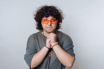 Person with curly hair wearing orange glasses and accessories poses with hands clasped against a...