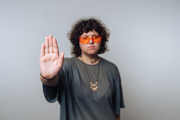 Individual with curly hair wearing orange sunglasses raises hand in a stop gesture against a plain...