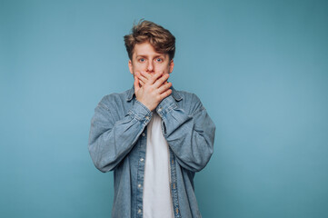 Young man covering mouth with hands against a light blue background showcasing surprise or shock