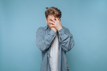 Young man expressing frustration while covering his face against a plain blue background during a...