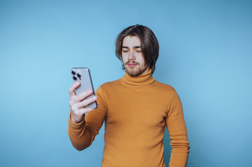 Young man wearing a turtleneck sweater looks at his smartphone in front of a solid blue background