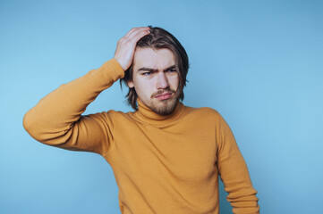 Young man expresses confusion while standing against a light blue background during indoor activity