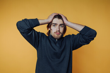 Frustrated young man with hands on head posing against a bright yellow background during a...