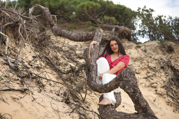 Young, beautiful, brunette woman, with red shirt and white pants, with sweet and tender look, sitting on a fallen log in the sand. Concept beauty, relax, nature, calm, happiness.