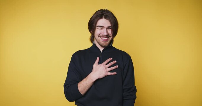 cheerful young man places his hand on his heart while smiling warmly, embodying warmth and sincerity on yellow background