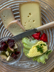 cheese pieces, pickled vegetables, butter and fruit in glass platter on wood table, Swiss rustic alpine hut look, top down view, vertical shot