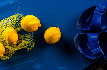 Lemons, sandals and net bag against a deep blue background. A vibrant composition featuring yellow lemons, net bag, and blue sandals set against a blue backdrop. Summertime vacation Well-being concept