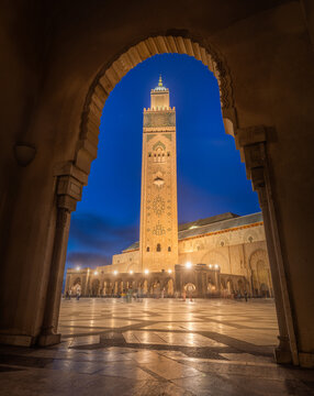 View of the Hassan II Mosque's minaret piercing the twilight sky, framed by a stone archway, casting a golden glow on the wet ground, Casablanca, Casablanca-Settat, Morocco.