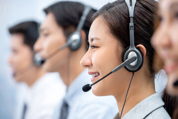 Asian woman telemarketer wearing headset with microphone working with colleagues in call center...