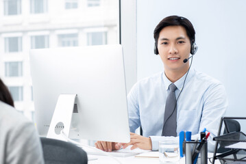 Smiling handsome Asian man wearing microphone headset working in call center office as a...