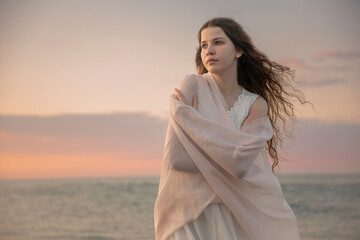 Young woman wrapped in a soft pink shawl, standing by the sea at sunset, gazing thoughtfully into...