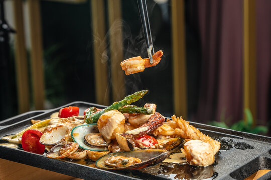 Chopsticks lift a steaming shrimp from a sizzling seafood platter featuring mussels, scallops, vegetables, and creamy sauce on a hot grill plate