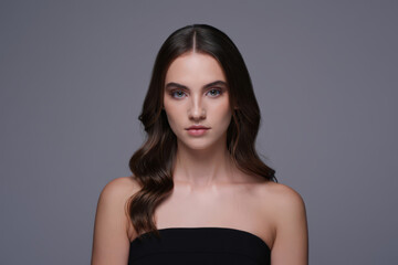 Naklejka premium Striking Portrait of a Brunette Woman with Wavy Hair and Bare Shoulders Against a Neutral Gray Background Emphasizing Natural Beauty and Contemporary Aesthetics