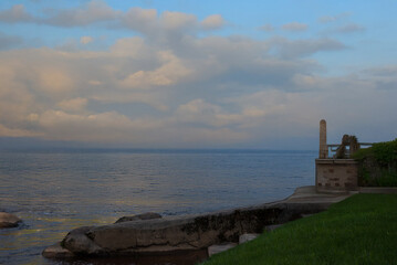 Fototapeta premium Calm sea view at sunset with dramatic clouds, a stone terrace, and lush grass in the foreground, creating a peaceful coastal atmosphere.