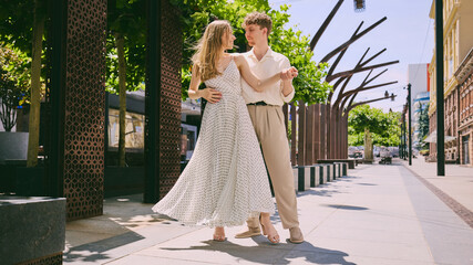 Young romantic couple dancing joyfully on sunny city street in elegant light summer outfits....