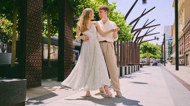 Young romantic couple dancing joyfully on sunny city street in elegant light summer outfits. Concept of love, lifestyle, romance, fashion, travel, happiness, and urban relationships.