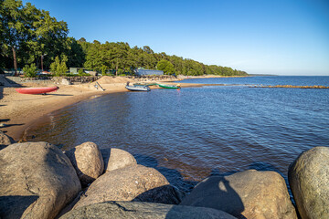 Sunny coast in Komarovo with round boulders and sandy beach. Blue water reflects sky, mixed forest...