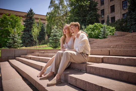 Pause and play. Couple sits closely on outdoor steps under a bright sunny day near lush green trees. Concept of summer romance and nature appreciation.