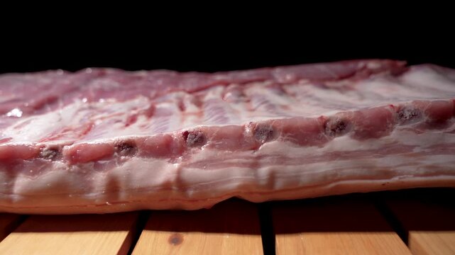 A large piece of raw pork with a rib structure lies on a wooden board. The meat is fatty and juicy, and the camera slowly pans along it.