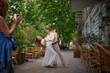 Couple twirling romantically in outdoor cafe setting as woman in jeans claps with cheerful support....