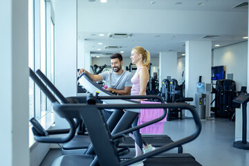 Mid adult woman and personal fitness trainer preparing for workout on treadmill at gym.