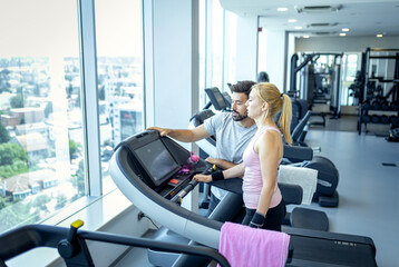 Mid adult woman and personal fitness trainer preparing for workout on treadmill at gym.
