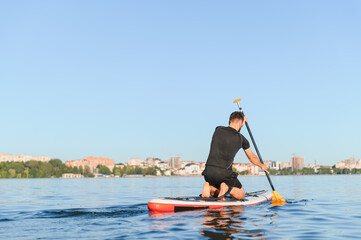 Kneeling athlete paddling on sup board on calm lake with cityscape in background