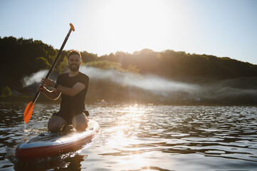 Sportsman kneeling on sup board paddling on lake at sunset