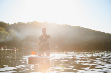 Man kneeling on sup board paddling on lake at sunset