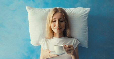 Woman waking up after sleeping on white pillow imagining she lying on bed holds cup of coffee